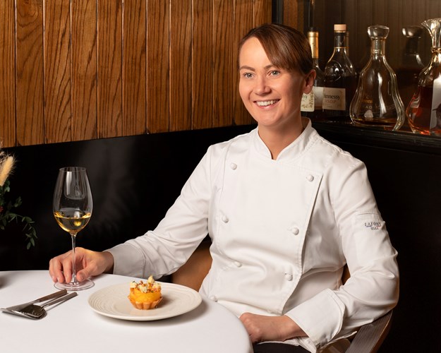 A woman sitting at a table with a glass of wine and a cake wearing a Chef's outfit