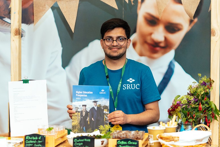 A student ambassador holding a booklet, behind a stall with food samples